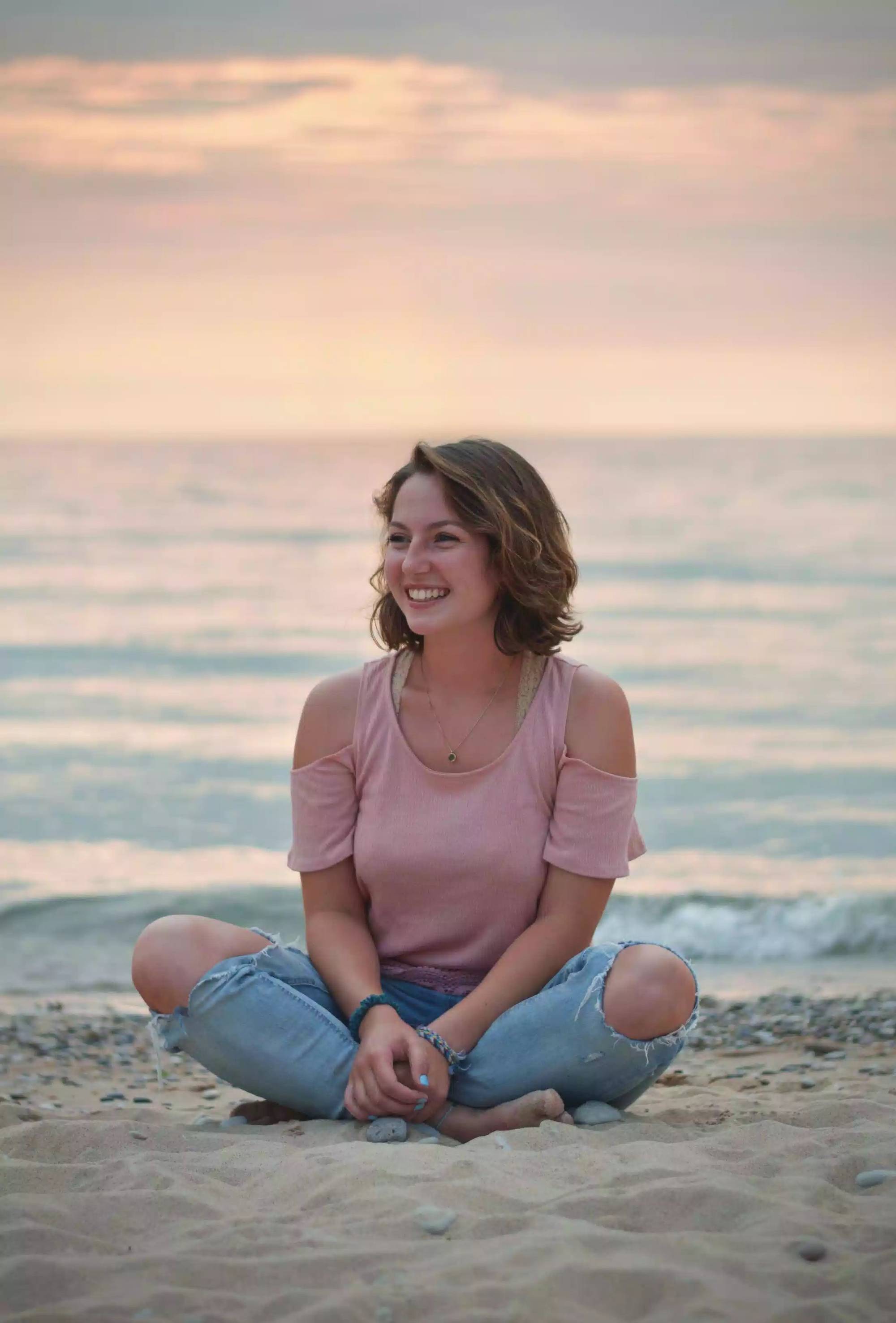 Girl sitting in the sand with a sunset behind her. She is wearing a pink t-shirt and blue jeans and smiling while facing to the left.